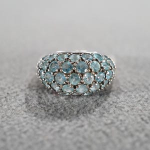 May include: A silver-toned ring featuring a dome of small, round, light blue gemstones. The ring's band is smooth, and the gemstones are closely set, creating a textured surface. The ring is set against a gray background.