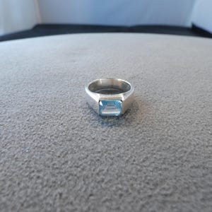 May include: A silver-toned ring featuring a rectangular, light blue faceted gemstone set in the center. The band is smooth and solid, tapering slightly toward the setting. The design is simple and clean, highlighting the contrast between the metallic silver band and the clear, pale blue stone.