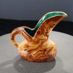 May include: A small, decorative pitcher with a rounded base and a curved spout. The pitcher is primarily brown with a marbled pattern, and the interior of the spout is green. It has a small handle and is likely ceramic.