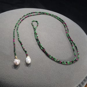 May include: A beaded necklace featuring green and red gemstones. The necklace has a lariat style with two white, teardrop-shaped pearls hanging from the ends. The necklace is displayed on a gray surface.