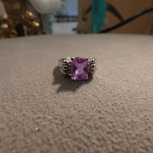 May include: A ring featuring a rectangular, faceted pink gemstone set in a silver-toned metal band. The setting includes small, gold-toned bead accents surrounding the central stone, creating a two-tone metallic appearance on the ring's band.