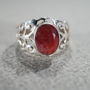May include: A silver ring with an oval, faceted, red gemstone. The ring has an openwork design around the gemstone. The ring is set against a neutral background.