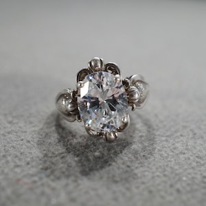 May include: A silver ring with a large, oval-cut, clear gemstone as the centerpiece. The ring has an ornate design with decorative elements around the gemstone and on the band. The ring is set against a gray background.