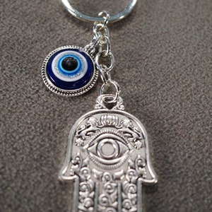 May include: Silver keychain with a blue evil eye charm and a silver hamsa hand charm. Both charms feature an eye design.