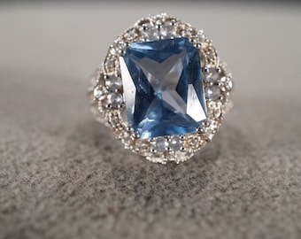 May include: A silver ring with a large, square, blue gemstone surrounded by smaller, clear gemstones.