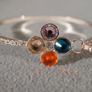 May include: A silver bangle bracelet with a row of clear rhinestones and a charm featuring five colorful gemstones. The charm is in the shape of a flower with a blue, orange, pink, yellow, and clear gemstone.