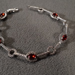 May include: A silver bracelet with red gemstones set in a circle design. The bracelet has a delicate chain and is adorned with small, sparkling stones.