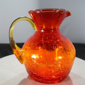 May include: A small, orange glass pitcher with a textured surface and a yellow handle. The pitcher is a vintage style.