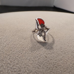 May include: A silver-toned ring with a unique design. The ring features two stylized leaf-shaped elements, one red and one white, with intricate floral detailing. The ring is set against a neutral background.