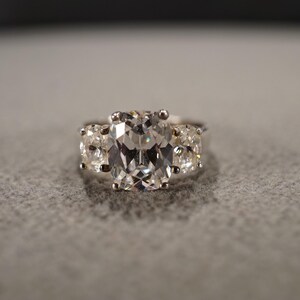 May include: A silver ring with three clear gemstones. The center stone is a large oval cut, and the two side stones are smaller round cuts.