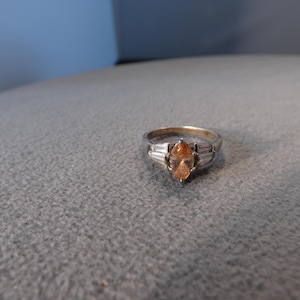 May include: A silver-toned ring featuring a central marquise-cut orange gemstone. The center stone is flanked on each side by three small, clear baguette-cut stones set into the metal band, creating a structured and symmetrical design.
