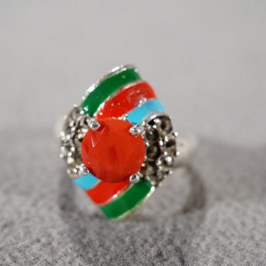 May include: A silver ring with a red, green, and blue enamel design. The ring has a large, round, red stone in the center.