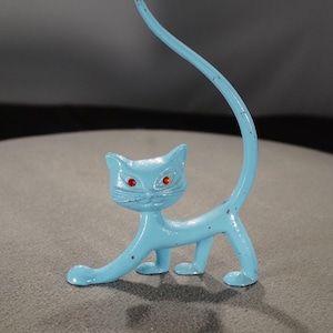 Puede incluir: Figura de gato azul claro con ojos rojos y cola arqueada. El gato está representado en posición de caminar, con una pata delantera extendida. La figura de metal tiene un acabado envejecido, lo que le da un aspecto vintage.