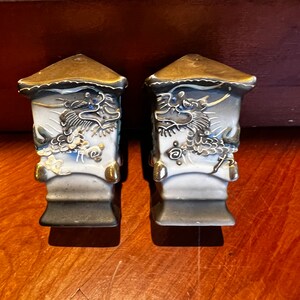 May include: A pair of ceramic salt and pepper shakers with a dragon design. The shakers are white with gold accents and blue and black details. The dragon design is on both sides of the shakers.