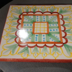 May include: A square ceramic tile with a colorful geometric design. The tile features a central square with a grid pattern surrounded by a border of green leaves and yellow accents.