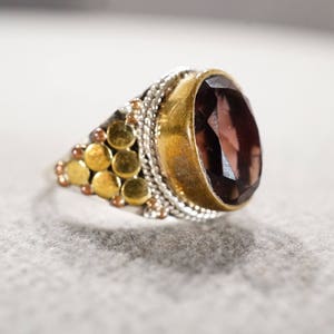 May include: A silver ring with a large, oval-shaped, dark purple gemstone set in a gold bezel. The ring band is decorated with a pattern of small, gold circles.