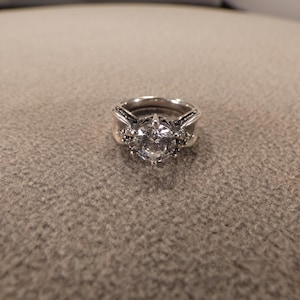 May include: A silver ring with a large, clear, round gemstone as the focal point. The ring has a detailed band with smaller accent stones. The ring is set against a neutral, textured background.