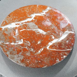 May include: A round, polished stone with a striking pattern of red, white, and gray veining. The stone is set against a soft, gray background. The overall effect is one of natural beauty and elegance.
