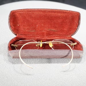 May include: Vintage rimless eyeglasses with gold-tone metal accents, resting inside an open, brown leather case with a red velvet interior. The glasses have a delicate, wire-frame design.