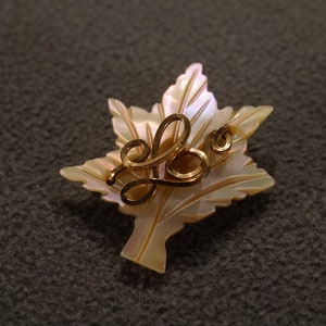May include: A gold leaf-shaped brooch with a delicate gold design in the center. The brooch is made of mother-of-pearl and has a pearlescent finish.