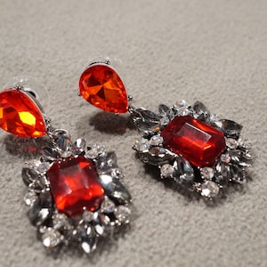 May include: A pair of dangle earrings with red crystal stones and silver-toned metal accents. The earrings feature a large, rectangular red stone surrounded by smaller, clear stones and a smaller, teardrop-shaped red stone.