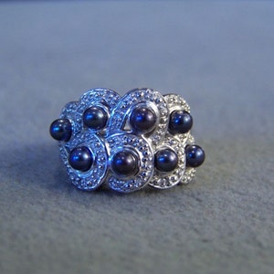 May include: A silver ring with a design of interlocking circles set with black pearls. The ring is made of sterling silver and features a polished finish.