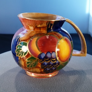 May include: A round ceramic pitcher with a gold handle. The pitcher is decorated with a colorful fruit design, including peaches, grapes, and a pear. The background is a mix of brown, blue, and gold.