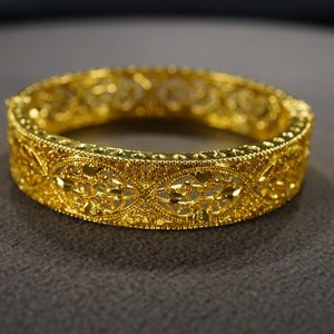 May include: A gold bangle bracelet with a delicate, intricate design. The bracelet is made of a thin, filigree metal and has a rounded shape.