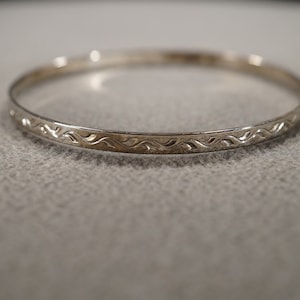 May include: A silver bangle bracelet with a repeating wave pattern. The bracelet is thin and circular, with the decorative pattern etched along the outer surface. The jewelry is displayed on a neutral gray surface.