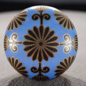 May include: A blue ceramic knob with a black and gold geometric design. The knob has a round shape and a smooth surface.
