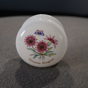 May include: White ceramic knob with a floral design featuring pink and white flowers, green leaves, and a blue butterfly. The text "Gazania Ringens" is written in gold lettering.