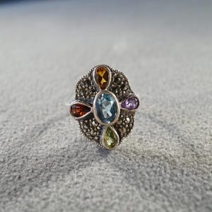 May include: A silver ring featuring a central oval blue gemstone surrounded by four smaller pear-shaped stones in orange, red, purple, and green. The setting is decorated with numerous small, dark, faceted marcasite stones in a floral pattern.