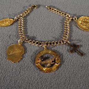 May include: A gold charm bracelet with a variety of charms, including a cross, a bird, a tree, and a profile of a person with the word "Teresa" inscribed.