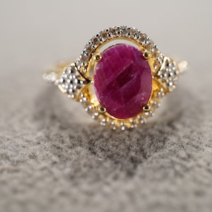 May include: A gold ring with a large oval ruby gemstone surrounded by smaller white gemstones.