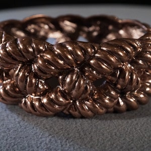 May include: A copper-colored ring with a rope-like design. The ring is made of a thick, braided metal and has a smooth, polished finish.
