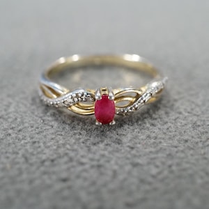 May include: A gold and silver-toned ring with a red oval-shaped gemstone at the center. The band features a twisted design with small, clear accent stones. The ring is set against a gray background.