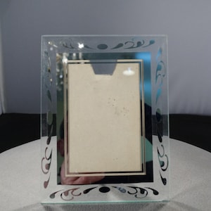May include: A clear glass picture frame with a mirrored surface and a decorative etched design. The frame has a black border and a white mat.