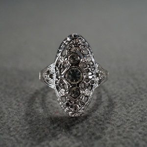 May include: Silver filigree ring with an oval design. The ring features three central clear stones and smaller accent stones. The intricate openwork design is visible throughout the ring's surface.