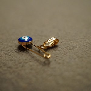 May include: A gold safety pin with a blue and white evil eye charm attached to it.