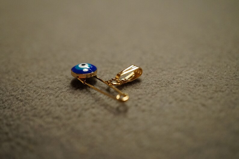 May include: A gold safety pin with a blue and white evil eye charm attached to it.