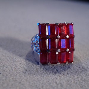May include: A silver ring with a large square red gemstone set in the center. The gemstone is surrounded by smaller silver accents.