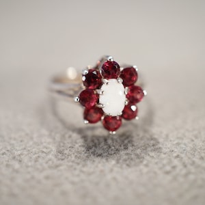 May include: A silver ring with a white opal center stone surrounded by a cluster of red gemstones.