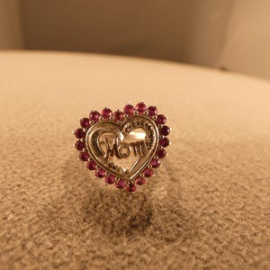 May include: A silver-toned ring in the shape of a heart, with the word "Mom" in the center. The heart is surrounded by small, round, red gemstones. The ring is set against a neutral background.