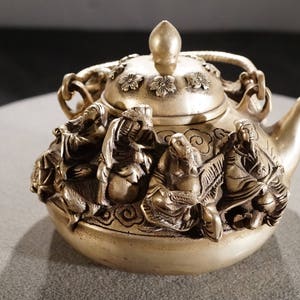 May include: A silver teapot with a lid and a spout, decorated with intricate carvings of figures and floral designs.