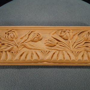 May include: A terracotta tile with a raised design of tulips. The tile is rectangular and has a smooth, slightly textured surface. The tulips are carved in detail, with their stems and leaves extending out from the center of the tile.
