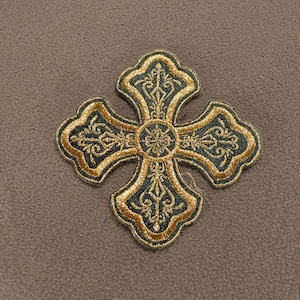 May include: A cross-shaped embroidered patch featuring a dark green fabric base with intricate gold thread detailing. The design includes ornate scrollwork patterns within each arm of the cross and a central circular motif with gold stitching.
