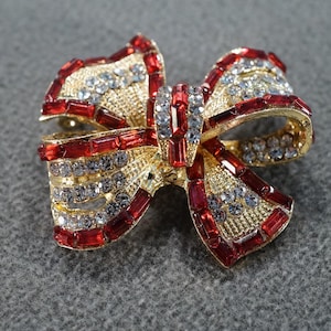 May include: Gold-tone bow brooch with red and clear rhinestones. The bow is tied in a knot with the ends of the bow pointing up.