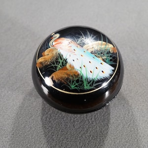 May include: A round, black decorative box with a painted peacock design. The peacock has a white and blue tail, with brown and gold accents. The box has a gold rim.