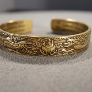 May include: A gold-toned metal bracelet with an intricate design of two dragons facing each other, surrounded by clouds. The bracelet is open and ready to be worn.