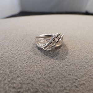 May include: A silver-toned ring featuring a crossover design with multiple bands. The center band is embellished with a row of small, clear, round-cut stones, while the surrounding bands have a smooth, polished finish and intricate detailing.